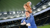 A dad carrying his young daughter on his shoulders. The daughter is throwing a soccer ball. There is a soccers stadium in the background. A dad carrying his young daughter on his shoulders. The daughter is throwing a soccer ball. There is a soccers stadium in the background.
