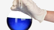 A gloved hand holding a flask with a blue liquid in front of a white background. A gloved hand holding a flask with a blue liquid in front of a white background.