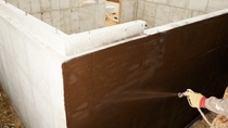 A worker is spraying waterproof sealant on a new home's concrete foundation. This area of the foundation will be below ground and the tar like material prevents water from contacting the concrete. Focus is on the wall in the area of the spray, the hand and sprayer are slightly soft.