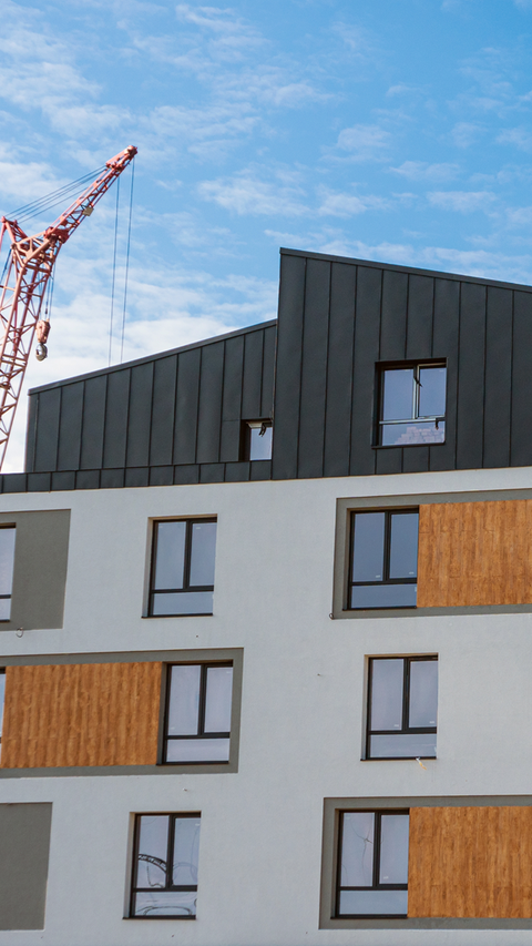 Modern apartment building under construction with a red crane to the left. The building has a black roof and wooden accents against a blue sky.
